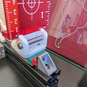 May include: A white and silver metal bracket with a red and green plastic piece attached to it. The bracket is mounted on a black metal track. A red ruler with a bullseye target is visible in the background.