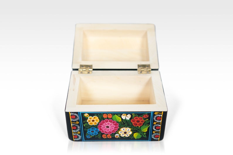 May include: Open wooden box with a black exterior and floral design. The box is rectangular with a hinged lid and brass-colored hinges. The floral design includes pink, red, yellow, and blue flowers with green leaves. The interior of the box is light-colored wood.