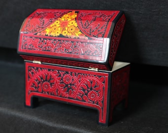 May include: A red wooden box with intricate floral and bird designs. The box is open and has a black interior. The box is on a black surface.