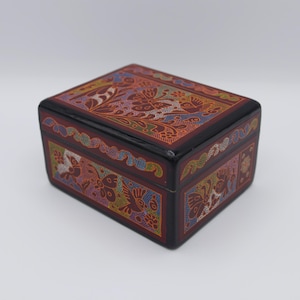 Könnte beinhalten: Eine rechteckige, dekorative Box mit dunkelroter Basis und schwarzem Rand. Die Box ist mit aufwendigen, farbenfrohen Mustern mit floralen und Vogelmotiven verziert. Das Design wiederholt sich auf Deckel und Seiten.