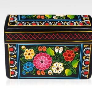 May include: Black wooden box with a curved lid, hand-painted with colorful floral designs. The box features a vibrant array of flowers in pink, yellow, blue, and red, with green leaves and white accents. The edges are decorated with geometric patterns.
