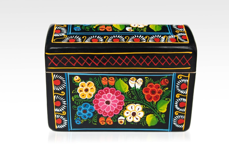 May include: Black wooden box with a curved lid, hand-painted with colorful floral designs. The box features a vibrant array of flowers in pink, yellow, blue, and red, with green leaves and white accents. The edges are decorated with geometric patterns.