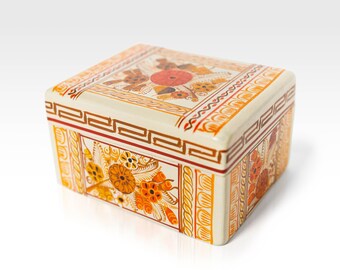 May include: A small, rectangular box with a white background and intricate orange and yellow floral designs. The box has a decorative border with a repeating geometric pattern.