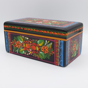 Könnte beinhalten: Eine schwarze Holzkiste mit aufwendigen handbemalten Blumendesigns in Rot, Orange, Gelb, Grün und Blau. Die Kiste ist rechteckig und hat einen Deckel mit Scharnier.