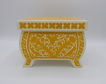 May include: A white decorative box with a yellow floral design and a geometric pattern on the top and bottom. The box has four legs.