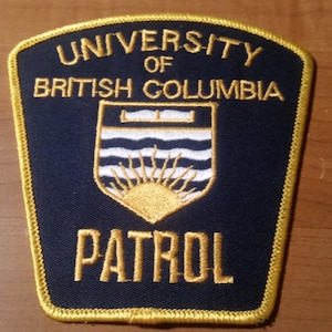 May include: A navy blue patch with gold embroidered text and trim. The text reads "UNIVERSITY OF BRITISH COLUMBIA" and "PATROL". A crest in the centre features a sunburst and waves.
