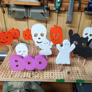 May include: A collection of wooden Halloween-themed cutouts on sticks, including orange pumpkins, white skulls, white ghosts, a black cat, a black bat, and a purple sign with the word BOO, all arranged upright on a perforated wooden board.