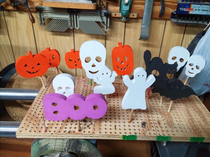 May include: A collection of wooden Halloween-themed cutouts on sticks, including orange pumpkins, white skulls, white ghosts, a black cat, a black bat, and a purple sign with the word BOO, all arranged upright on a perforated wooden board.