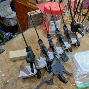 May include: A series of black plastic trigger clamps securing several small, heart-shaped wooden pieces. The clamps feature long, thin metal bars and are arranged in a row. The wooden pieces have a light-colored, textured surface on one side.