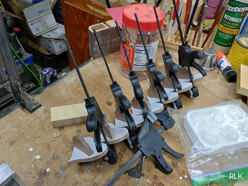 May include: A series of black plastic trigger clamps securing several small, heart-shaped wooden pieces. The clamps feature long, thin metal bars and are arranged in a row. The wooden pieces have a light-colored, textured surface on one side.