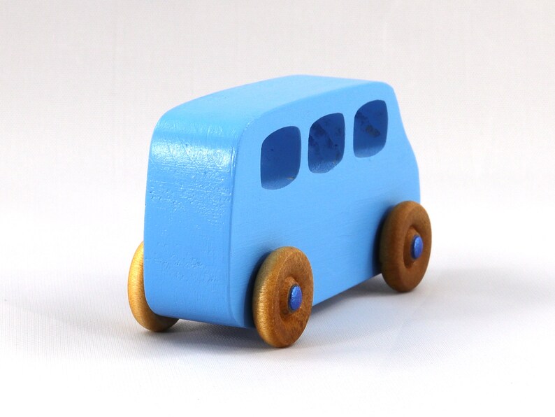 May include: A light blue wooden toy bus with three windows and four brown wheels. The bus is painted a light blue color and has a simple design.
