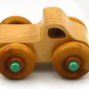 A handmade wooden toy monster truck with a natural wood body and four large wheels. The body and wheels are finished with amber shellac, and the hubs are painted with metallac emerald green acrylic paint. The toy is designed with a simple, classic aesthetic.