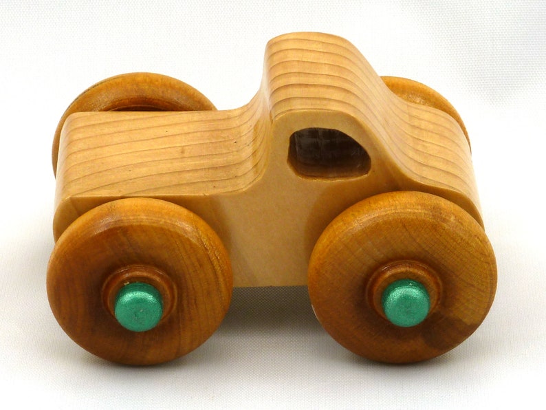 A handmade wooden toy monster truck with a natural wood body and four large wheels. The body and wheels are finished with amber shellac, and the hubs are painted with metallac emerald green acrylic paint. The toy is designed with a simple, classic aesthetic.