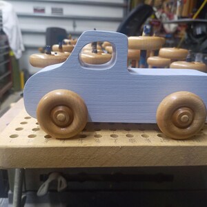Side view of a pale blue wooden toy pickup truck with a cutout cab window and two amber-finished wooden wheels, photographed in the workshop.