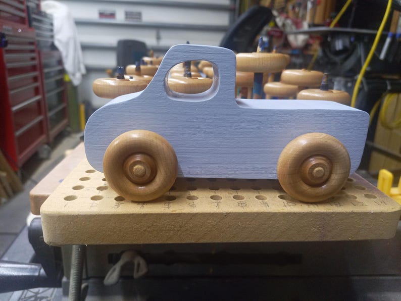 Side view of a pale blue wooden toy pickup truck with a cutout cab window and two amber-finished wooden wheels, photographed in the workshop.