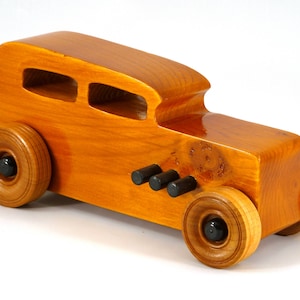 May include: A handcrafted wooden toy car with a classic design. The car is made of polished wood with a warm, amber tone. It features large wooden wheels with black centers and three black cylinders on the side. The car has two openings on the roof.