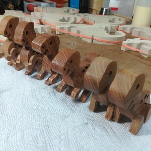Row of six handmade wooden baby dinosaur toys in different hardwoods on a toymaker’s workbench, with wooden parts and works in progress visible in the background.