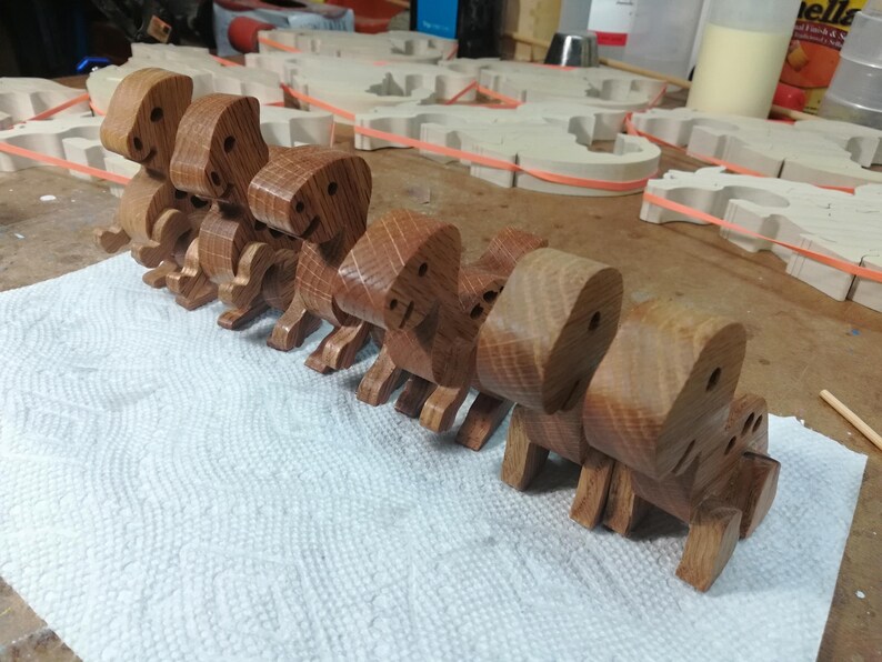 Row of six handmade wooden baby dinosaur toys in different hardwoods on a toymaker’s workbench, with wooden parts and works in progress visible in the background.
