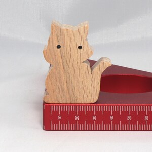 May include: A wooden cat figurine sits on a red, triangular ruler with a measurement scale in inches. The ruler has a circular cutout in the center.