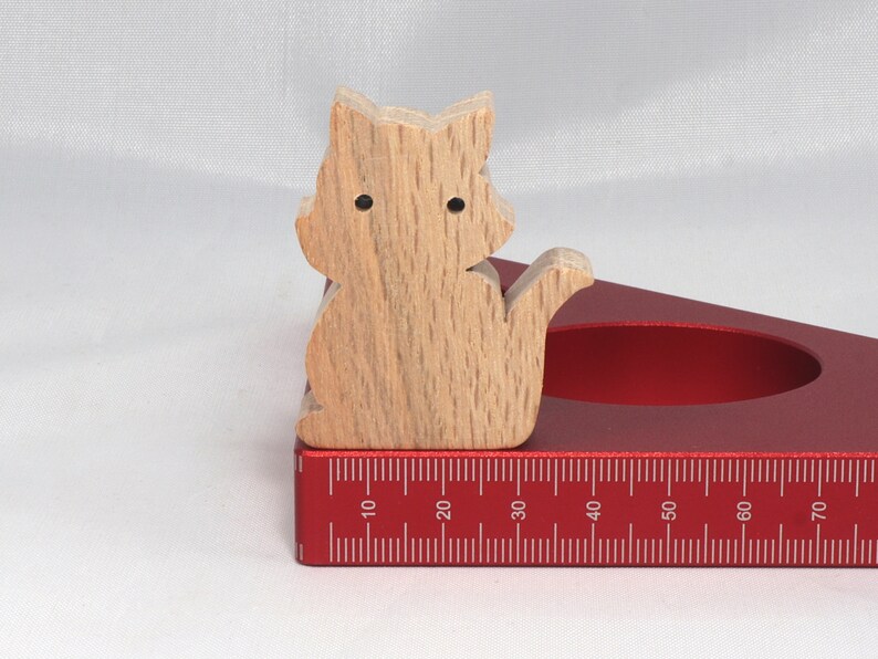 May include: A wooden cat figurine sits on a red, triangular ruler with a measurement scale in inches. The ruler has a circular cutout in the center.