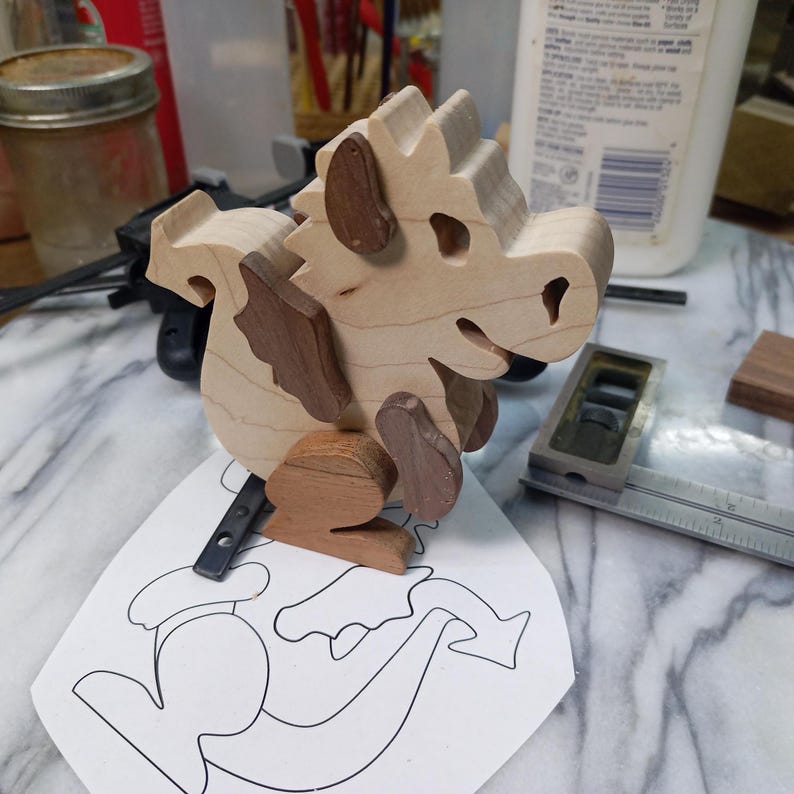 An unfinished wooden baby dragon with dark wings and feet stands on its paper pattern at the workbench.