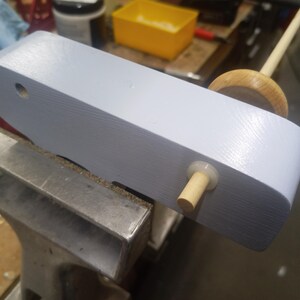 Close-up of the underside of a pale blue wooden toy truck held in a vise, showing a birch axle and nylon washer during wheel assembly.