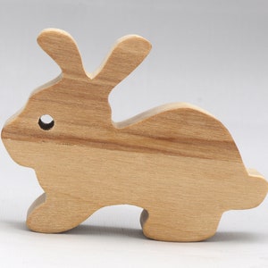 May include: A wooden rabbit cutout, painted in a natural wood color. The rabbit is lying down with its head turned to the side. The rabbit has a small hole for an eye.