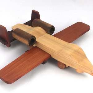 May include: A wooden toy airplane with a natural wood finish, featuring dark brown engine housings and wings, and reddish-brown wings and tail. The toy has small wooden wheels and is designed for play.