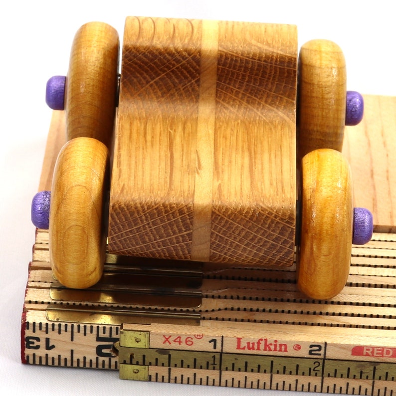 May include: A handcrafted wooden toy car with a natural wood finish. The car features four wooden wheels with purple accents and is mounted on a base made from a folding ruler. The car's body is made of light and dark wood.