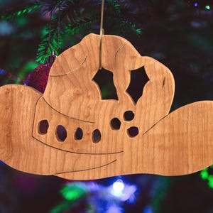 Handmade wooden Christmas tree ornament shaped like a cowboy hat, featuring a sprig of holly tucked into the hat band, star-shaped leaf cutouts, and round berry accents.