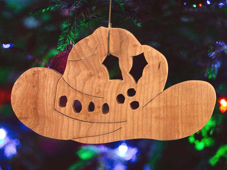 Handmade wooden Christmas tree ornament shaped like a cowboy hat, featuring a sprig of holly tucked into the hat band, star-shaped leaf cutouts, and round berry accents.