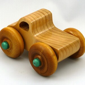 A handmade wooden toy monster truck with a natural wood body and four large wheels. The body and wheels are finished with amber shellac, and the hubs are painted with metallac emerald green acrylic paint. The toy is designed with a simple, classic aesthetic.