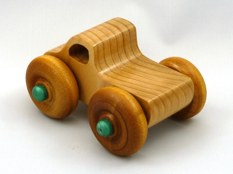 A handmade wooden toy monster truck with a natural wood body and four large wheels. The body and wheels are finished with amber shellac, and the hubs are painted with metallac emerald green acrylic paint. The toy is designed with a simple, classic aesthetic.