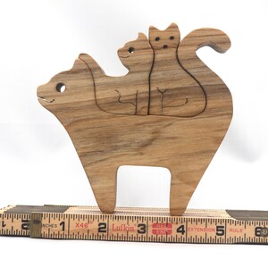 A freestanding wooden puzzle cut in the shape of a large cat standing on four legs. Two smaller kitten figures nest on the large cat’s back and interlock as puzzle pieces. Simple drilled holes form the eyes and nose on each cat, with a shallow cut line for the mouth on the large cat. The completed puzzle stands upright on a plain white surface. Handmade by Odin’s Toy Factory In Tallahassee, Florida, USA.