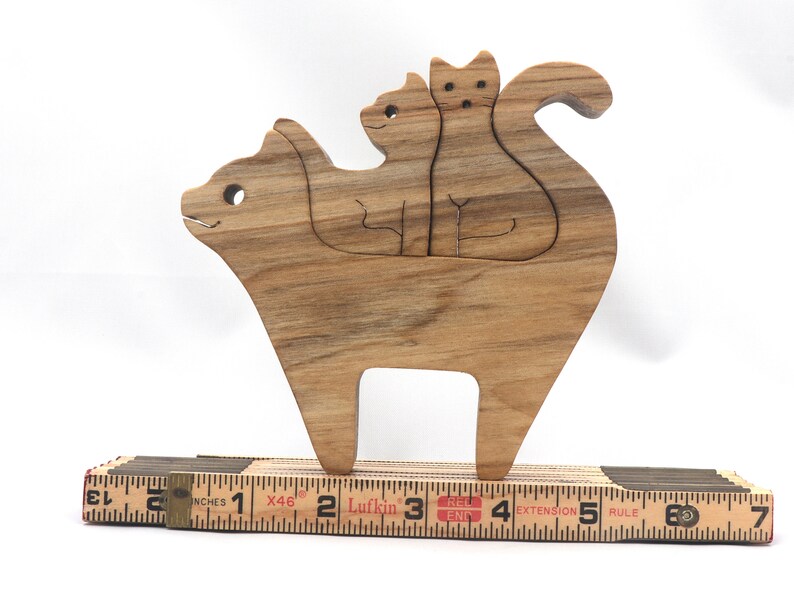 A freestanding wooden puzzle cut in the shape of a large cat standing on four legs. Two smaller kitten figures nest on the large cat’s back and interlock as puzzle pieces. Simple drilled holes form the eyes and nose on each cat, with a shallow cut line for the mouth on the large cat. The completed puzzle stands upright on a plain white surface. Handmade by Odin’s Toy Factory In Tallahassee, Florida, USA.