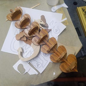 May include: A series of interconnected wooden heart-shaped shelves with a visible wood grain pattern. The structure is arranged in a linear formation, featuring small horizontal platforms attached to each heart. Paper templates marked with 1/2 inch are visible.
