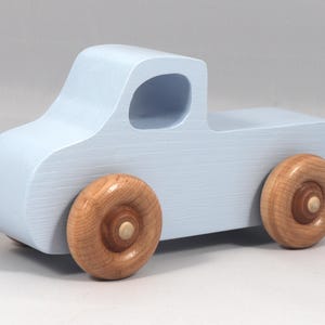 May include: A light blue wooden toy truck with natural wood wheels. The truck has a classic pickup design with a cab and a cargo bed. The wheels are round and attached to the truck's body. A simple and charming toy.