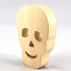 A handcrafted, unfinished light wood Halloween skull cutout featuring carefully carved eye sockets, a nose, and a cheerful, smiling mouth. Displayed upright against a white background.