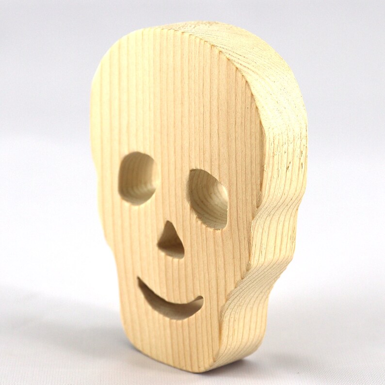 A handcrafted, unfinished light wood Halloween skull cutout featuring carefully carved eye sockets, a nose, and a cheerful, smiling mouth. Displayed upright against a white background.