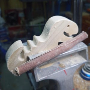 Unfinished wooden T-Rex body held in a bench vise during sanding, with sawdust on its edges and a sandpaper-wrapped sanding stick resting across the vise.