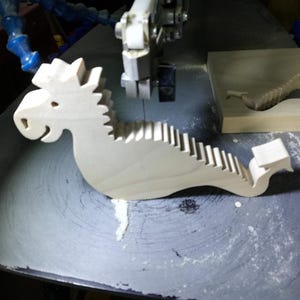 Freshly cut wooden baby dragon body resting on a scroll saw table beside the remaining hardwood blank and sawdust.
