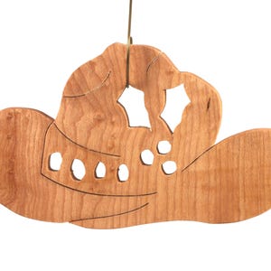 Handmade wooden Christmas tree ornament shaped like a cowboy hat, featuring a sprig of holly tucked into the hat band, star-shaped leaf cutouts, and round berry accents.