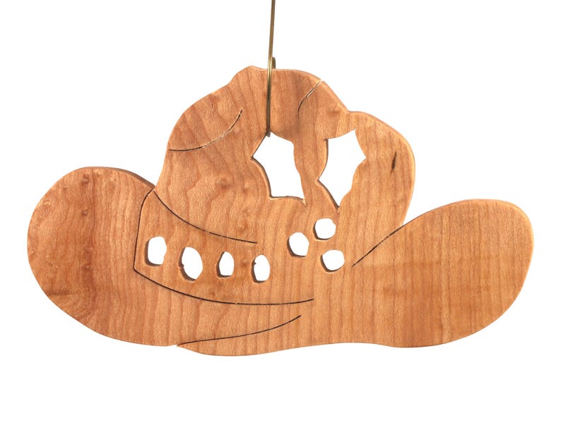 Handmade wooden Christmas tree ornament shaped like a cowboy hat, featuring a sprig of holly tucked into the hat band, star-shaped leaf cutouts, and round berry accents.