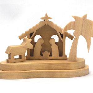 May include: Wooden nativity scene featuring a stable, figures of Mary, Joseph, and baby Jesus, a donkey, and a palm tree. The figures are carved from light-colored wood and set on a tiered base. A star sits atop the stable.