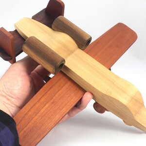 May include: A wooden toy airplane in shades of brown, tan, and dark brown. The toy is held in a person's hand. The plane has a propeller, wings, and a tail. The toy is made of wood and appears to be handmade.