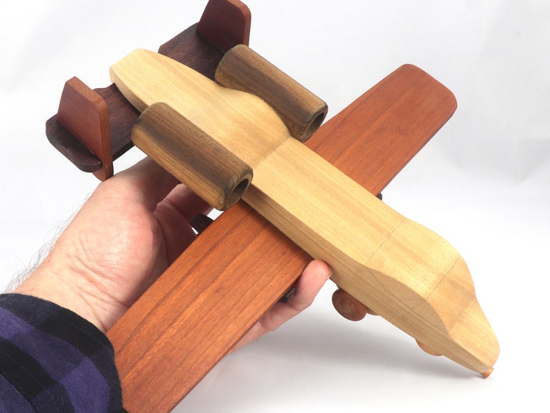 May include: A wooden toy airplane in shades of brown, tan, and dark brown. The toy is held in a person's hand. The plane has a propeller, wings, and a tail. The toy is made of wood and appears to be handmade.