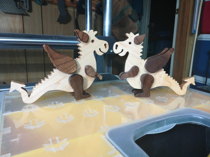 Two handmade wooden baby dragon figurines with light hardwood bodies and dark contrasting parts facing each other in the woodworking shop.