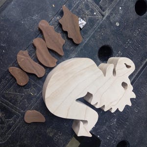 An unfinished light wood dragon body lies on a workbench beside several dark wood pieces cut for its wings and legs.