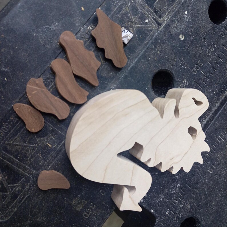 An unfinished light wood dragon body lies on a workbench beside several dark wood pieces cut for its wings and legs.