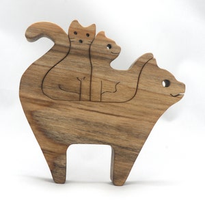 A freestanding wooden puzzle cut in the shape of a large cat standing on four legs. Two smaller kitten figures nest on the large cat’s back and interlock as puzzle pieces. Simple drilled holes form the eyes and nose on each cat, with a shallow cut line for the mouth on the large cat. The completed puzzle stands upright on a plain white surface. Handmade by Odin’s Toy Factory In Tallahassee, Florida, USA.